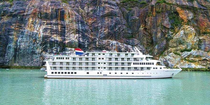 The American Constellation Coastal Cruise Ship sailing through the light green colored passage of Tracy Arm Fjord, surrounded by a cliff with bright gold and white streaks down the side.