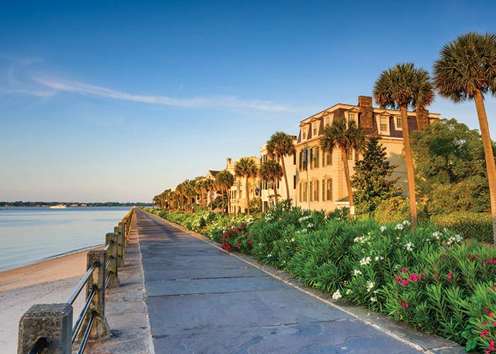 The Battery, a long strip of antebellum homes along the Charleston peninsula. There is a stone wall separating The Battery from the sand. In the distance, the blue sky is beginning to turn into a purple and orange sunset.