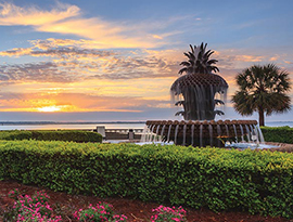 The Pineapple Fountain, a large fountain with water flowing down from layers that looks like a pineapple split in three sections, with the crown as the top layer in Charleston Waterfront Park. Perfectly trimmed bushes surround the circular pool below the fountain, and the Inland Passage and several palm trees are in the distance under a cloudy sunset.