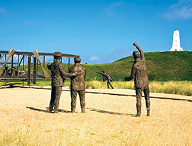 A recreation of the first flight, with a large sculpture of an airplane with a statue of a person laying on it, and four statues on the ground behind it at Wright Brothers National Memorial in Kitty Hawk.