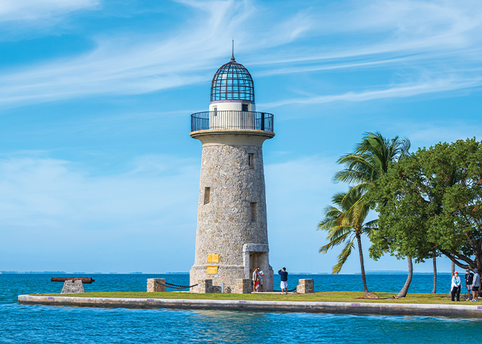 Boca Chita Key, a stone lighthouse with a wraparound black balcony at the top, almost completely surrounded by water besides a narrow path to land in Key Biscayne. There are a few palm trees in the foreground beside the lighthouse and a few tourists getting their picture taken in front of it.