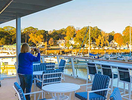 A guest on the sun deck of a Coastal Cat taking a picture with her phone of the boats docked along the shore and the full trees in the distance. The sundeck features several blue and white striped chairs and small white tables, and an American flag posted to the glass railing.