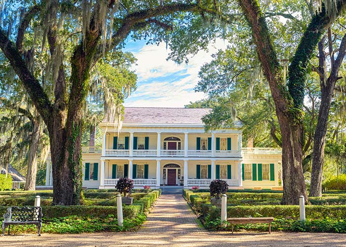 The historic St. Francisville landmark the Rosedown, a yellow house with bright green shutters and two-story white pillars with a brick pathway surrounded by perfectly trimmed bushes and large trees leading to the red door. 