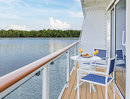 On an American Riverboat private balcony, two blue and white chairs surround a round table with a plate of food and two yellow drinks with large lemons on the rim on the balcony of one of the ships staterooms.