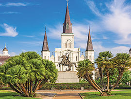 The St. Louis Cathedral with three spires stands in Jackson Square in New Orleans, while a statue of a rider on a horse is centrally placed in a manicured park with trees and bright greenery.