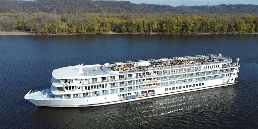 The American Riverboat American Serenade gliding along the Mississippi River, with a green landscape in the distance.