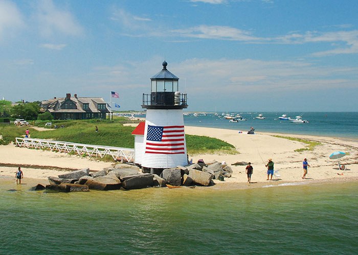 Brant Point Lighthouse, a small, white lighthouse on Nantucket Island with a large American flag on its side surrounded by rocks on the edge of the beach, along the water of Vineyard Sound. There is a ramp leading to the lighthouse from the grassy area behind the sand. People are fishing on the edge of the sand, and there are boats on the water and a large building in the middle of the island.