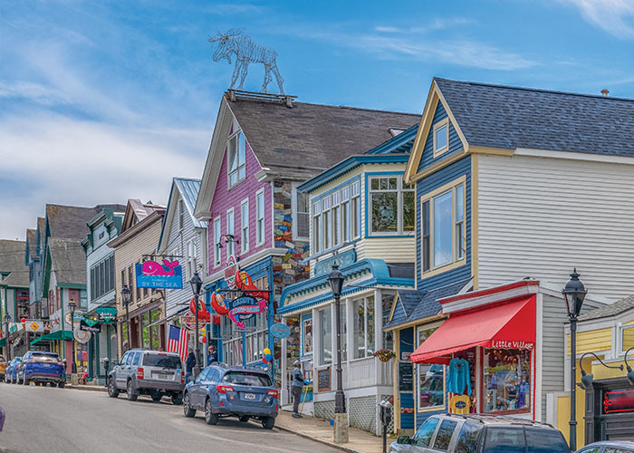 A roadside view of a line of buildings in Downtown Bar Harbor, with several cars parked alongside the sidewalk. One of the buildings is pink, one is yellow, and the others are a mixture of blue and tan.