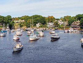 Dozens of jet boats and sail boats slide along the harbor in Ogunquit. There are several houses and trees along the shore in the background.