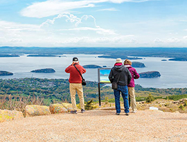 Three people stand along the edge of a cliff that is lined with large rocks looking over the harbor in Acadia National Park. One person is taking a photo of the water, while the others are looking at a map.