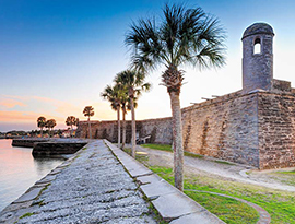 Castillo de San Marcos, a stone masonry fort in a grassy area, with a stone wall separating the land from the bay in St. Augustine. In the far distance, there are boats docked along the shore under a pink sunset that is reflecting on to the water.