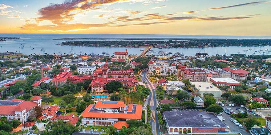 An aerial view of St. Augustine cityscape under an orange sunset over the distant bay. Most of the buildings have orange or red rooftops, creating a vibrant scene.