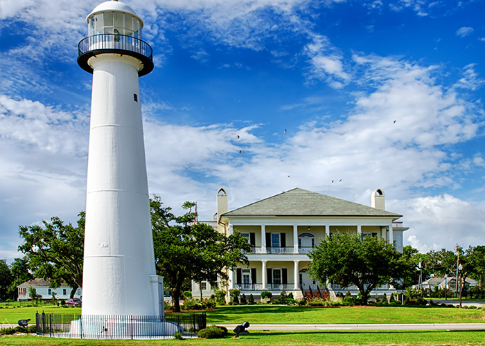 Biloxi Lighthouse, a tall white lighthouse with a small black fence around the bottom of it in Biloxi. There is a two-story yellow house with pillars on the front, bright green grass, and short trees in the background.