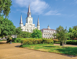 The St. Louis Cathedral with three dark grey spires stands in Jackson Square in New Orleans, while a statue of a rider on a horse is off to the right surrounded by bright green trees and bushes.