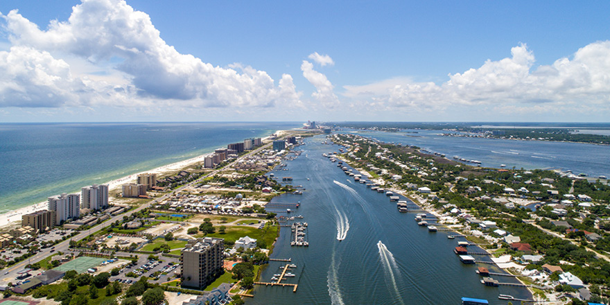 An aerial view of Pensacola, with the Bayou Texar dividing the two sides of the city, and Pensacola Bay in the distance. There are several boats sailing along Bayou and docked along the shore, where there are countless buildings and trees.