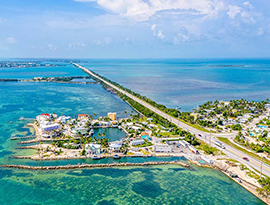 The Overseas Highway, a long stretch of road with trees on each side spanning far into the distance, surrounded by the bright blue ocean in Key West. Towards the closer portion of the highway, there is a small area of land on both sides that is occupied by trees and buildings.