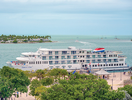 American Glory Coastal Cat docked on the bright blue water at Mallory Square in Key West. There are several trees in the foreground, and in the distance, there is a part of the city stretching around the water with a small boat gliding past.