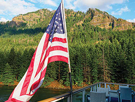 A vibrant American flag flying from the glass railing on the deck of an American Riverboat cruise ship while sailing along the Columbia River. Alongside the river, there is a cliff with thick forests spanning up the side. 