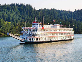 An American Cruise Lines paddlewheeler cruise ship sailing along the Columbia River. There are thick forests alongside the shoreline.