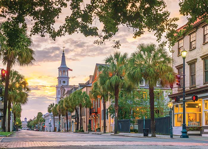 A line of both red brick and colorful buildings and tall palm trees along a street in Downtown Charleston, including the tall, white spire on top of St. Michael's church. 