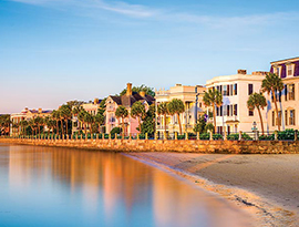 The Battery, a long strip of antebellum homes along the Charleston peninsula. There is a stone wall separating The Battery from the sand. In the distance, the blue sky is beginning to turn into a purple and orange sunset.