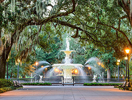 Forsyth Fountain, a tall white fountain with water spraying from the sides, surrounded by a short black metal fence and a wide pathway in Savannah. Live oak trees, lamp posts with an orange glow, and park benches line the pathway leading up to the fountain.