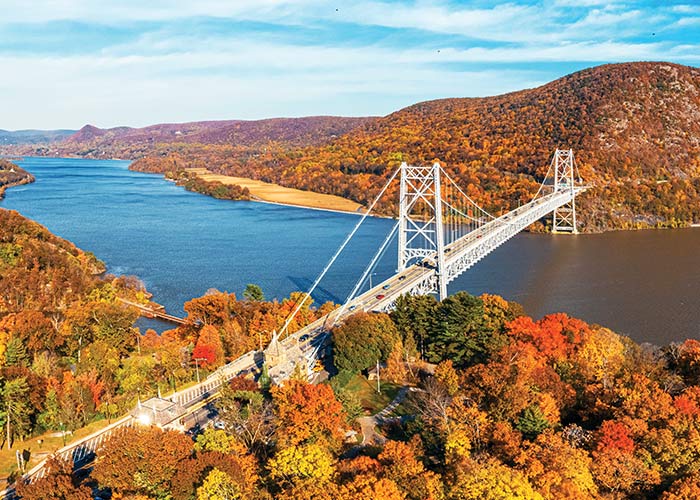 An aerial view of a long bridge across the Hudson River, and the hills covered in trees with vibrant red, orange, and yellow leaves on each side of the river during prime fall foliage season.