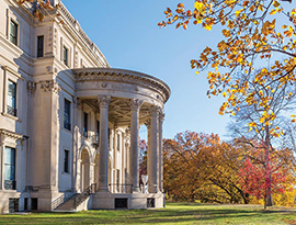 Vanderbilt Mansion National Historic Site in Hyde Park, a three-story, cream colored mansion with four tall pillars in front of the entryway stairs. There are several trees with orange and red leaves in the distance.