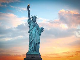 A close-up view of the Statue of Liberty, a blue-green colored statue of a woman with a crown on her head holding a torch in the air with fluffy, light pink clouds in the background.