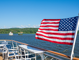 On the deck of an American Cruise Lines Coastal Cat, there is a large American flag, with its pole on the glass railing, blowing in the wind with the views of the Hudson River. There are several blue and white chairs and a two white tables on the deck. The Esopus Meadows Lighthouse is in the middle of the river.