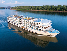 The American Riverboat American Serenade gliding along the Mississippi River, with the tree-filled shore and a bridge in the background. 