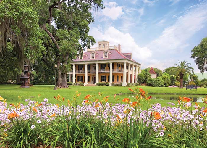 A historic plantation in Louisiana, Houmas House Plantation has two story pillars on all sides, grand porches and is set amongst beautiful grounds and gardens. There are pink, purple, and orange colored flowers in the foreground, and a small pond along the right side of the land.