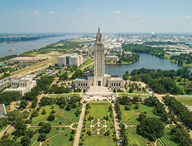 Aerial view of the Louisiana State Capitol building surrounded by landscaped gardens, trees, and a river winding through the Baton Rouge cityscape in the background. 