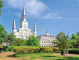 The St. Louis Cathedral with three dark grey spires stands in Jackson Square in New Orleans, surrounded by bright green trees and bushes.