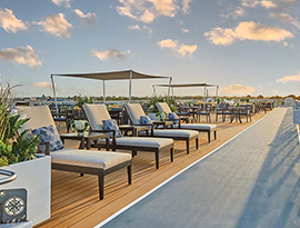 Several white lounge chairs with blue patterned pillows are placed next to a large plant in a grey, square pot along the sky walk on the American Riverboat sun deck.