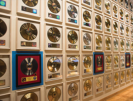 A wall of the Country Music Hall of Fame in Nashville, completely filled with dozens of gold and silver records in white frames. On the second row from the bottom, there are records with a red background and blue frame after every third frame.