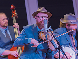 Three men are performing a live concert at the Grand Ole Opry in Nashville. The one man in focus in the middle is wearing a brown hat, circular glasses, and a blue suit. He is singing into a microphone while playing the violin. One of the men in the background is playing the cello, and the other is playing the banjo and also wearing the same hat.