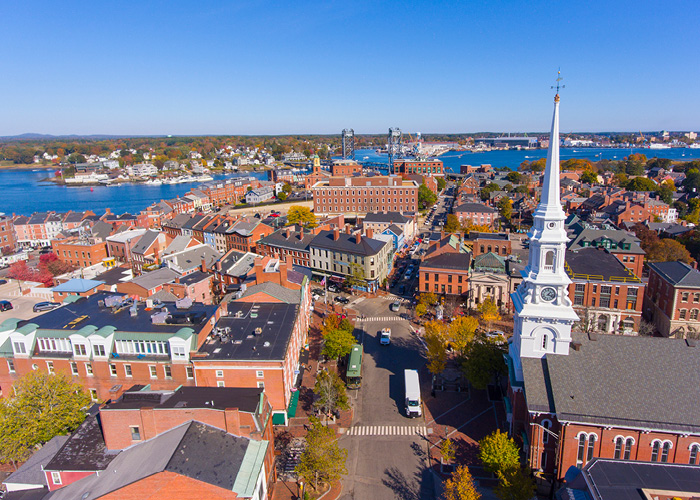 An aerial view of the Market Square in Portsmouth, where countless orange-tinted brick buildings fill the land, and the white spire from North Church stands tall above the other buildings. In the distance separating the city from the Maine horizon is the Piscataqua River.