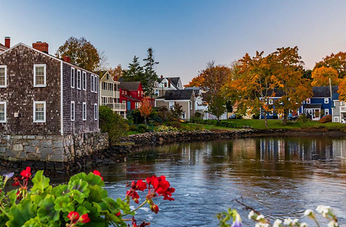 Several houses, including one that is completely supported by rocks, line the rocky edge of the harbor in Portsmouth. There are several trees with orange and red leaves on the shore.