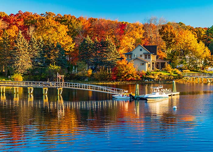 A small boat docked beside a long bridge on the Maine coast, with views of the autumn leaves on the full trees. There is a house on the shore that also has a long bridge leading out to the water.