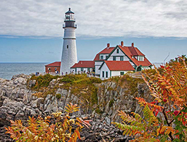 Portland Head Light, a white lighthouse next to a large white building with a red roof and green trim, on the edge of a rocky cliff beside Casco Bay. There are several people walking alongside the lighthouse.