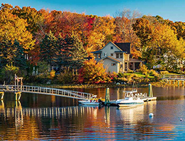 A small boat docked beside a long bridge on the Maine coast, with views of the autumn leaves on the full trees. There is a house on the shore that also has a long bridge leading out to the water.