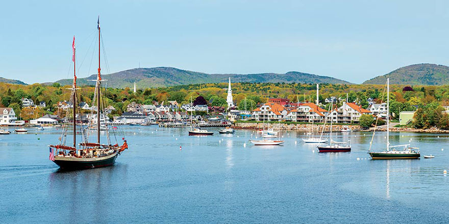 A view of several boats docked along the harbor in Camden, with small mountains and colorful fall foliage trees in the distance.