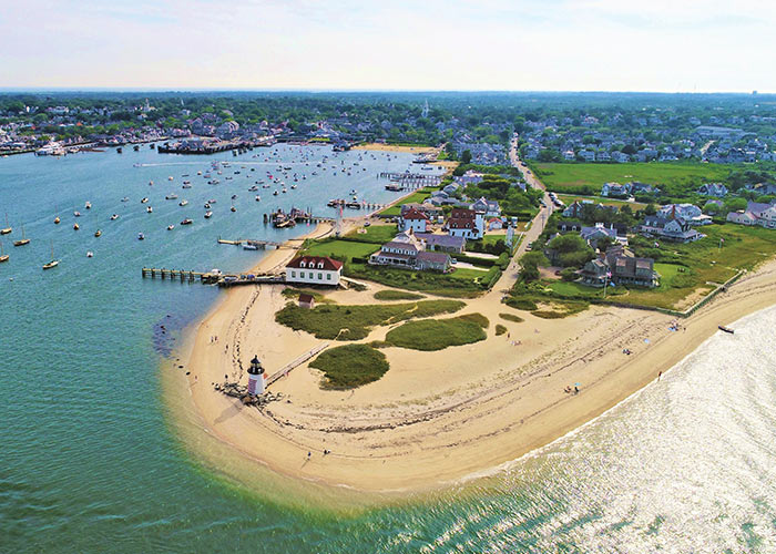 An aerial view of Nantucket Island, where the sand curves out into the water creating a U-shape. Brant Point Lighthouse is on the edge of the sand, and there are several buildings along the grassy parts of the island, and many boats docked along the water.