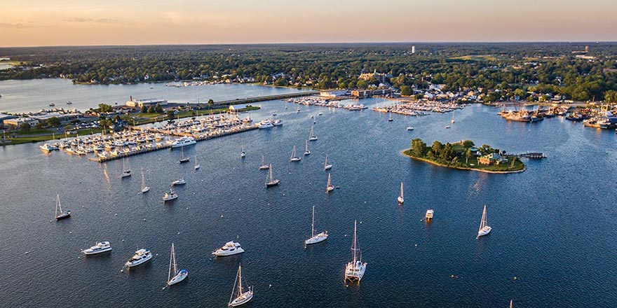 An aerial view of the bay with countless sailboats docked alongside an island and several sailing along the water with the city of New Bedford in the distance. 