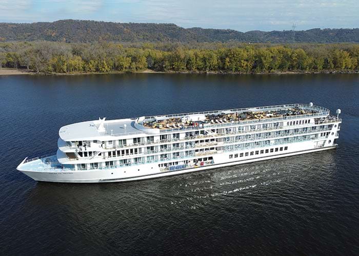 American Melody, an American Riverboat small cruise ship gliding along the Mississippi River, surrounded by lush greenery on the nearby shore.