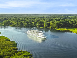A white American Cruise Lines American Riverboat small cruise ship navigates the Mississippi River, surrounded by lush, green forests under a clear sky.