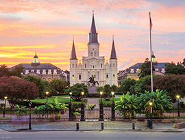 The St. Louis Cathedral with three spires stands in the background of Jackson Square in New Orleans, while a statue of a rider on a horse is centrally placed in a manicured park with trees and lampposts with an orange and pink sunset.