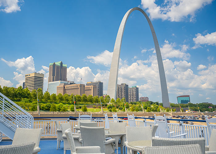 Multiple tables and chairs line the blue American Riverboat deck floor with the view of the St. Louis Gateway Arch and several skyscrapers in the background.