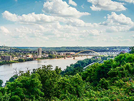 Aerial view of the Ohio River with a bright green nature scene in front, and a large city in the distance. 